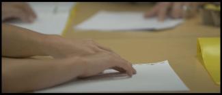 student hands reading braille on page with teacher's fingers across the table on far side of table.
